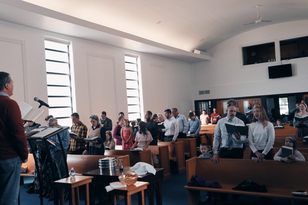 Sunday worship at Christ Church Livingston County: a leader reads from the lectern with the Lord's Supper table set in the foreground while the congregation stands and sings